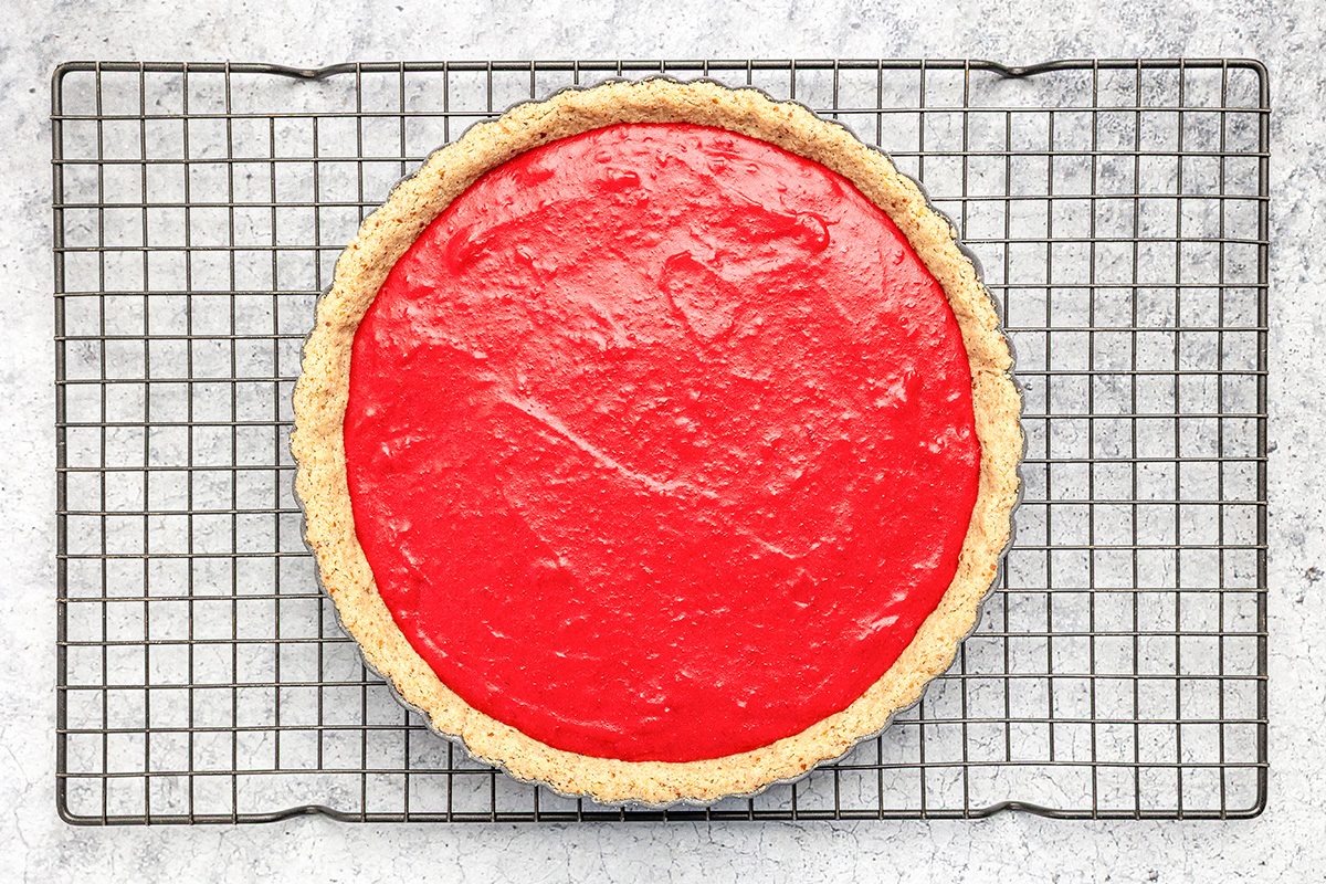 A round pie with a bright red filling sits on a cooling rack over a light gray, textured surface. The pie has a golden, crumbly crust and a smooth, glossy top.