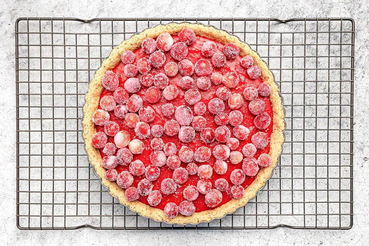 A tart topped with red and pink cherries sits on a cooling rack over a light, textured surface. The tart has a golden crust and the cherries appear fresh and glossy.