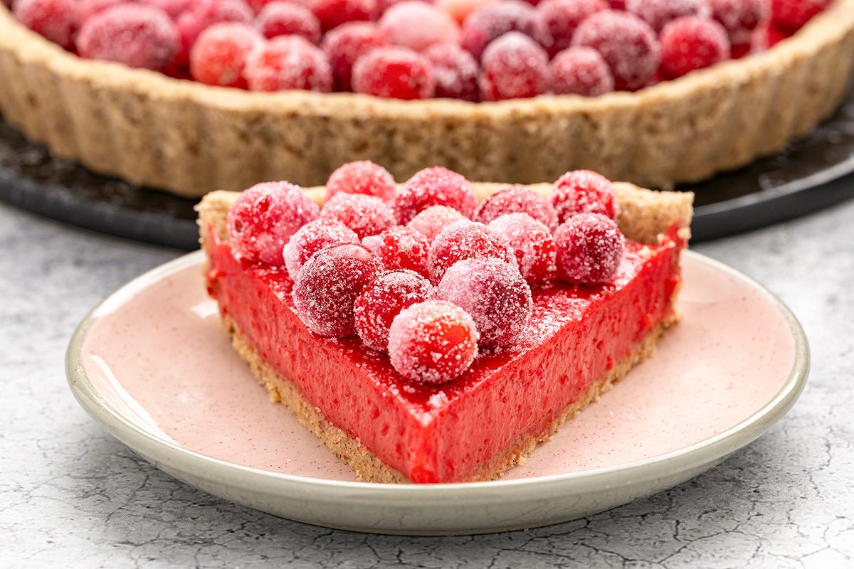 A slice of bright red cranberry tart with a crumbly crust, topped with sugared cranberries, sits on a pink plate with the rest of the tart visible in the background.
