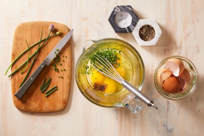 A whisk rests in a glass bowl containing eggs, chopped herbs, salt, and pepper. Nearby are a cutting board with chives and a knife, salt and pepper containers, and a glass jar with eggshells.