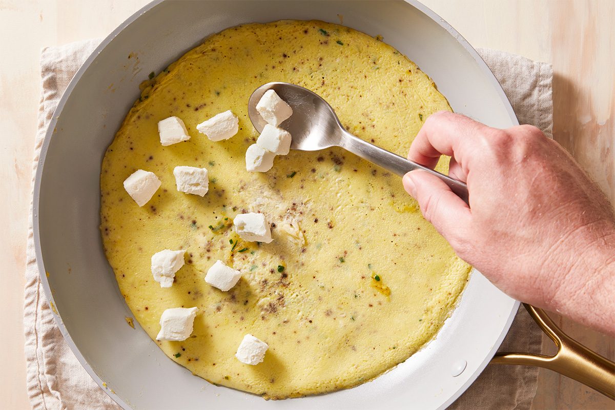 A hand uses a spoon to add small cubes of white cheese onto an omelette cooking in a frying pan. The pan sits on a beige cloth on a light-colored surface.