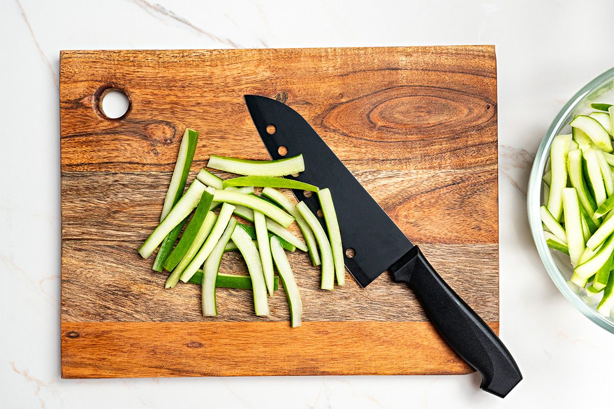 A pile of cucumber sticks and a black kitchen knife are on a wooden cutting board; a glass bowl with more cucumber sticks is partially visible to the right.