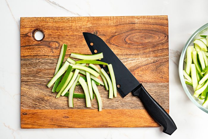A pile of cucumber sticks and a black kitchen knife are on a wooden cutting board; a glass bowl with more cucumber sticks is partially visible to the right.