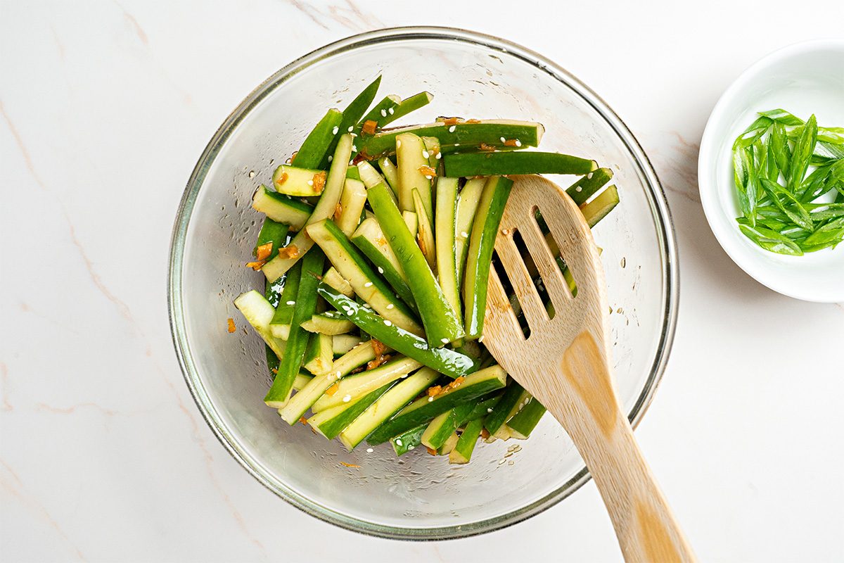 A glass bowl filled with sliced cucumbers mixed with seasonings sits on a white surface. A wooden slotted spoon rests in the bowl, and a small white dish with sliced green onions is nearby.