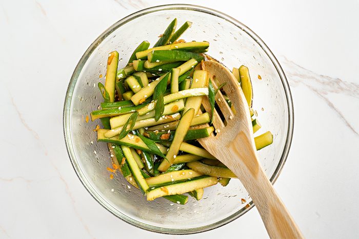 A glass bowl filled with thinly sliced cucumber sticks mixed with seasonings, next to a wooden serving fork and spoon, on a white marble surface.