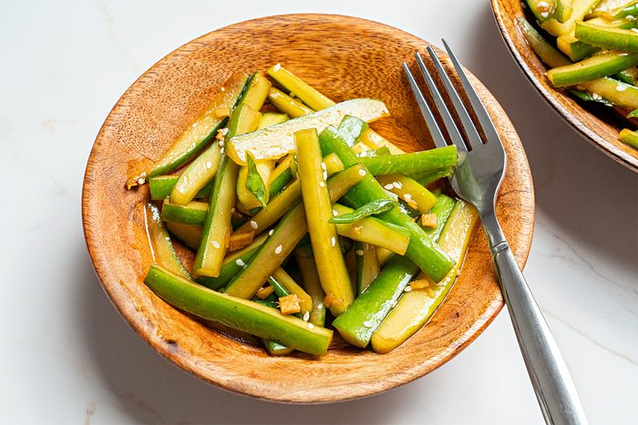A wooden bowl filled with sliced cucumber salad, garnished with sesame seeds and a light dressing. A fork rests on the edge of the bowl, all placed on a white surface.