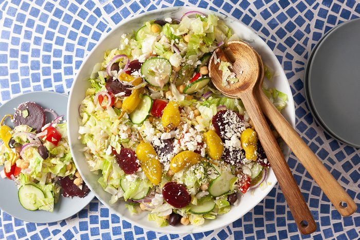 A large bowl of salad with cucumbers, beets, olives, mandarin oranges, feta cheese, lettuce, and red onions, with wooden salad servers, sits on a blue patterned table next to a small serving plate and an empty plate.