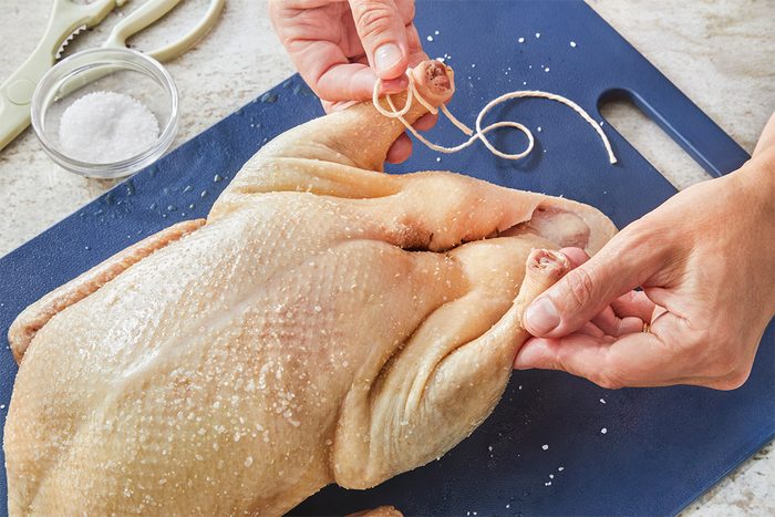 A person ties the legs of a raw, salted whole chicken with kitchen twine on a blue cutting board; kitchen scissors and a small bowl of salt are nearby.