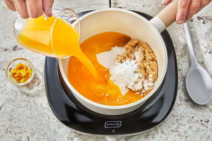 A person pours orange juice from a glass into a saucepan containing brown sugar and white powder, on a black electric stove. A small bowl of orange zest and a spoon are nearby on the countertop.