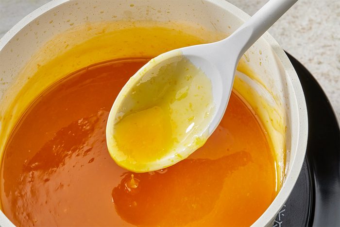 A white saucepan filled with bright orange and yellow melted sugar mixture, with a white spoon resting inside, coated in the thick, glossy liquid.
