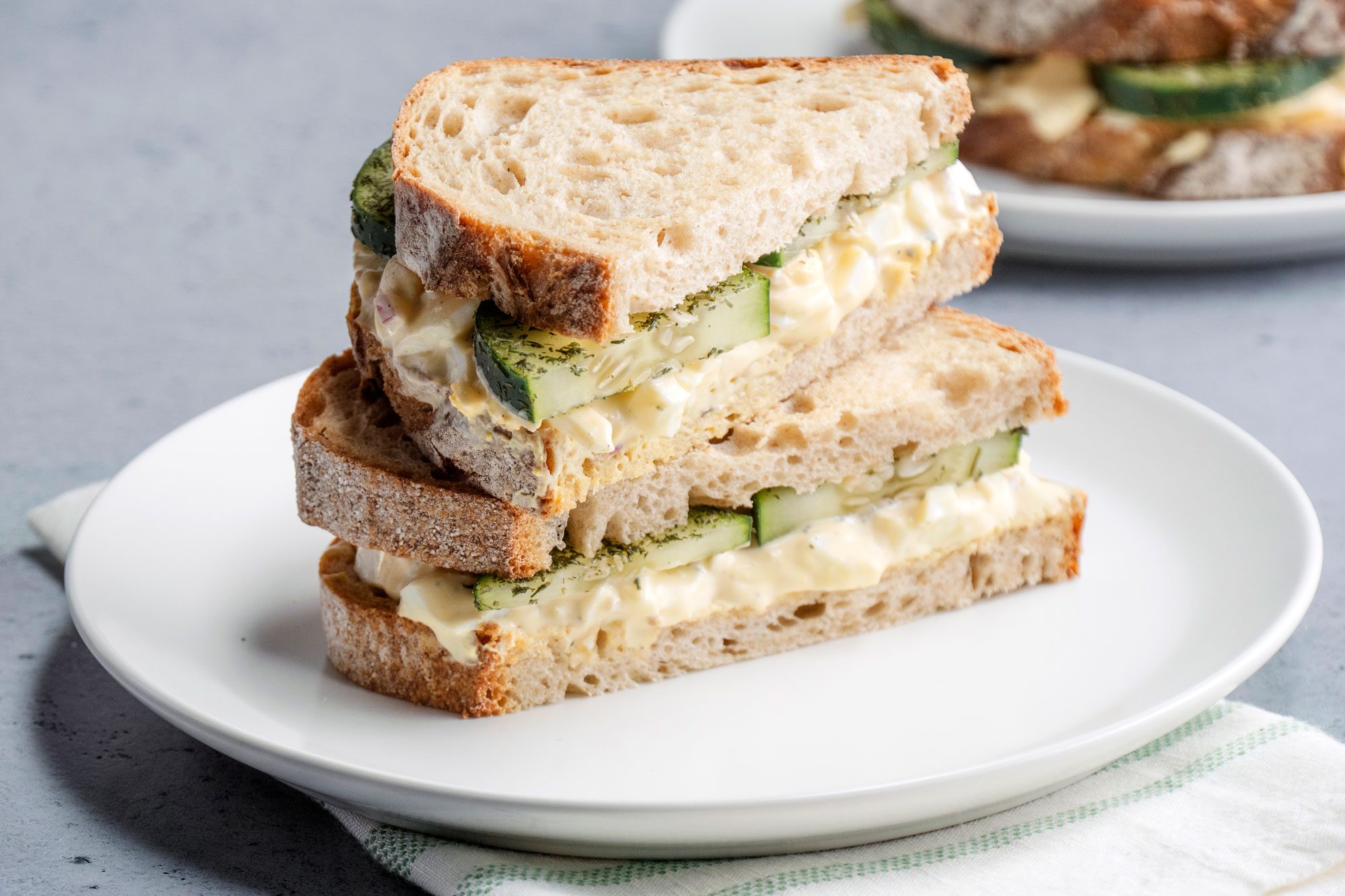 Close-up shot of Egg Salad and Cucumber Sandwiches served on two plates; one plate placed over a white napkin, the other in the background; all set on a grey surface