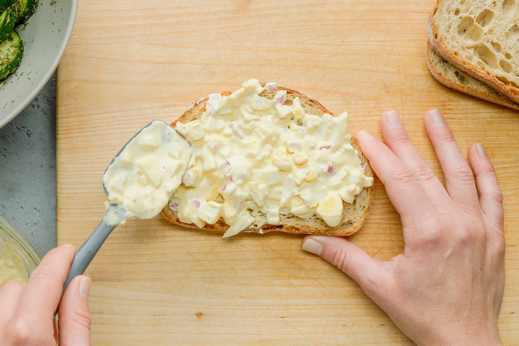 Overhead shot of egg salad spread over slices of toast, arranged on a wooden board; all set on a grey surface