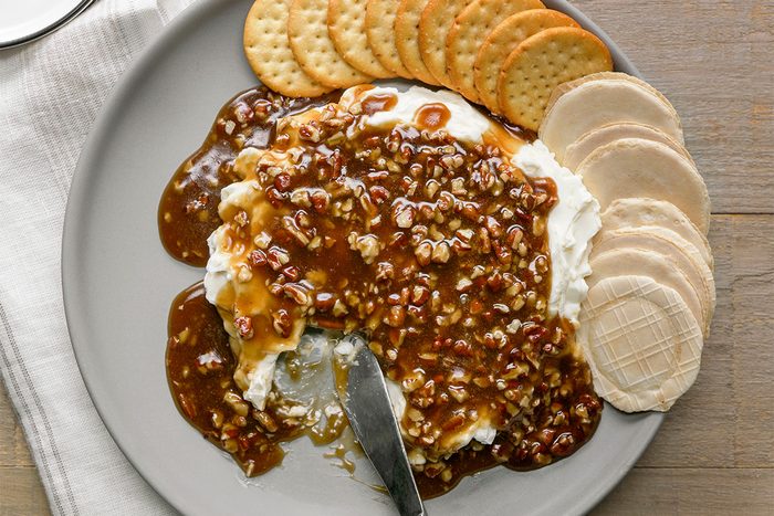 A plate with cream cheese topped with a brown, nutty sauce, surrounded by round crackers. A knife rests on the plate, partially in the cheese spread.