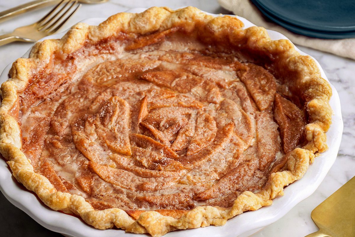 A freshly baked apple pie with a golden, crimped crust and thinly sliced apples arranged in a spiral pattern, sprinkled with cinnamon and sugar, sits in a white pie dish on a marble surface.