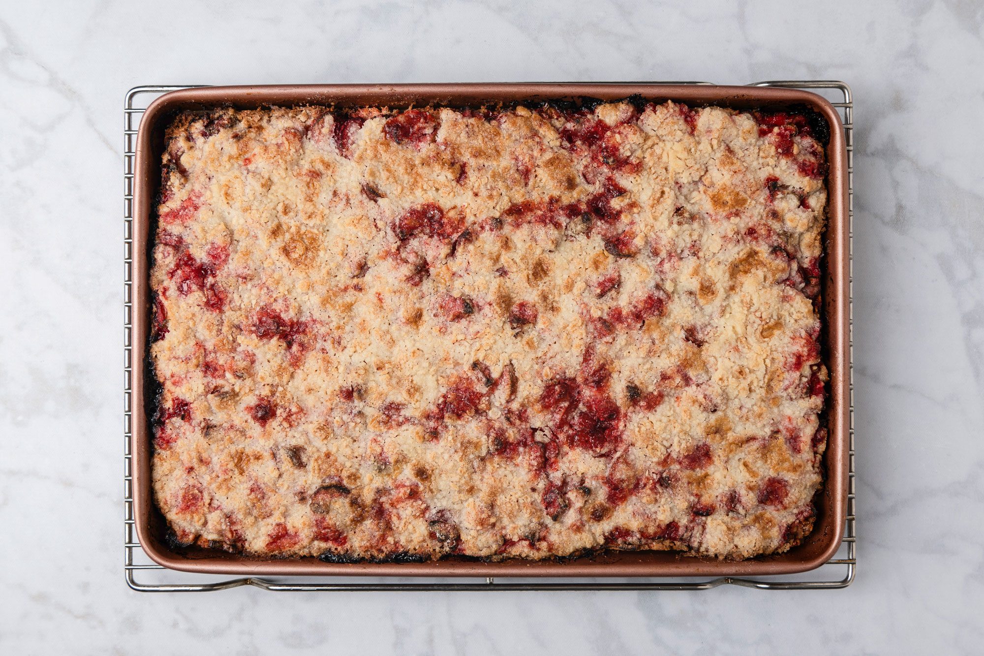 overhead shot of a rectangular baking pan filled with a golden brown, crumbly dessert topped with red fruit, possibly a berry crumble or crisp, sits on a wire rack on a white marble surface