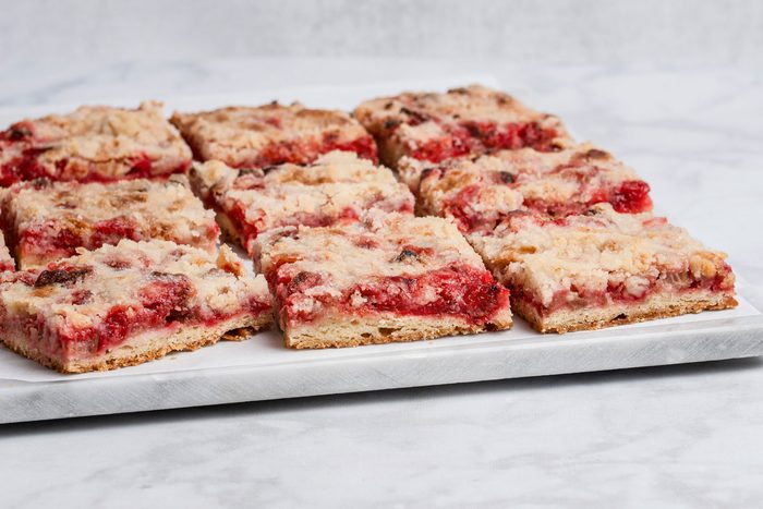 closeup left tilted shot of Nine rectangular Gluten Free Rhubarb Bars with a red filling and crumbly topping are arranged on a white marble surface, with a light grey, textured background