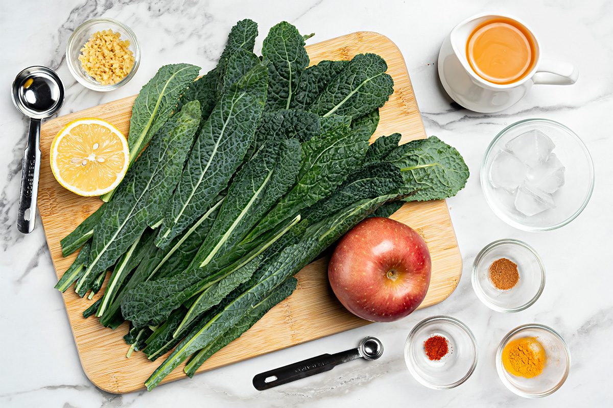 A wooden cutting board with fresh kale leaves, half a lemon, and an apple, surrounded by small bowls of spices, ginger, and a cup of coffee on a marble countertop.