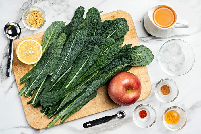A wooden cutting board with fresh kale leaves, half a lemon, and an apple, surrounded by small bowls of spices, ginger, and a cup of coffee on a marble countertop.
