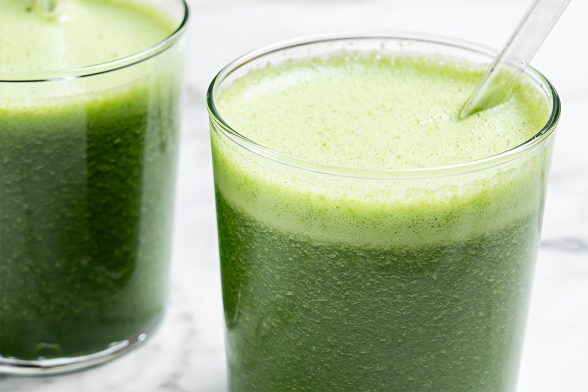 Two clear glasses filled with a frothy, vibrant green smoothie or juice, with one glass showing a metal straw. The drink rests on a white surface.