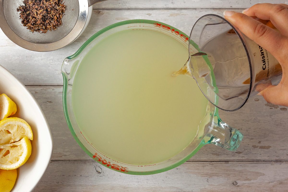 Top Shot of a person pours liquid from a measuring cup into a glass jug of cloudy yellow lemonade; sliced lemons sit in a bowl; tea leaves rest in a strainer on a white wooden surface