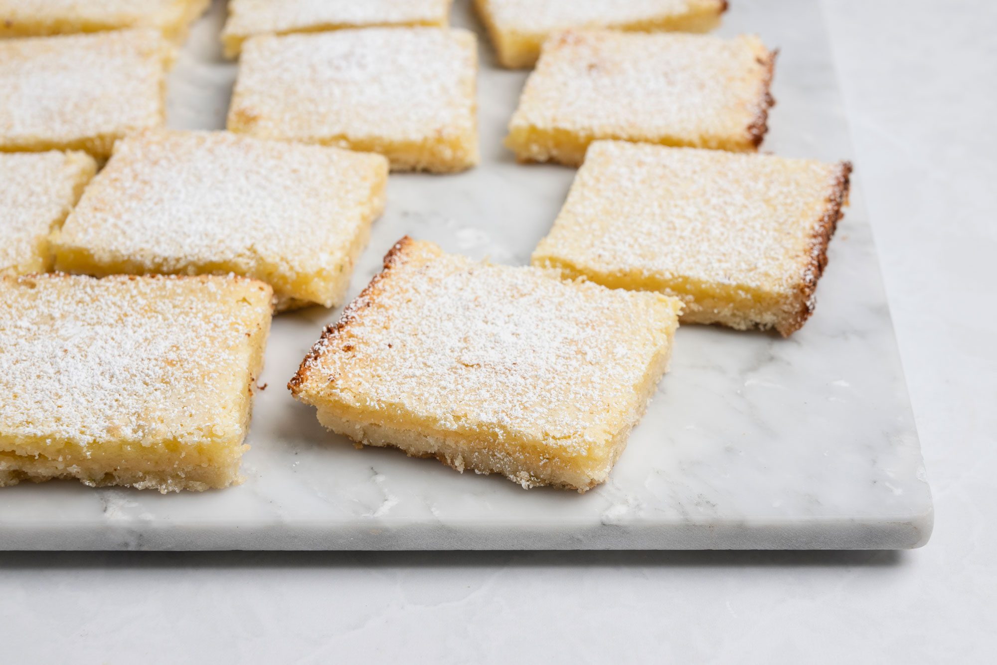 A 3/4th shot of a Neatly arranged lemon bars with golden edges and light yellow filling rest on a white marble background; each square is generously dusted with powdered sugar, creating an inviting and fresh presentation