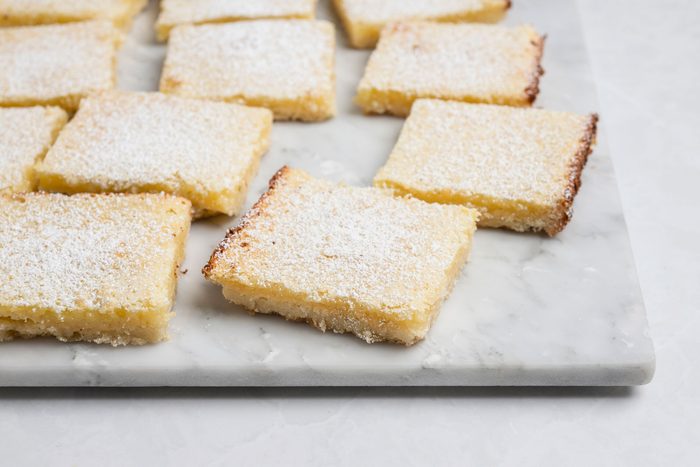 A 3/4th shot of a Neatly arranged lemon bars with golden edges and light yellow filling rest on a white marble background; each square is generously dusted with powdered sugar, creating an inviting and fresh presentation