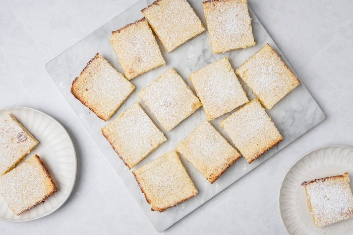 Top shot of Two lemon bars dusted with powdered sugar rest on a white plate; additional bars and a fork appear blurred in the background; bright lighting enhances the dessert's yellow color