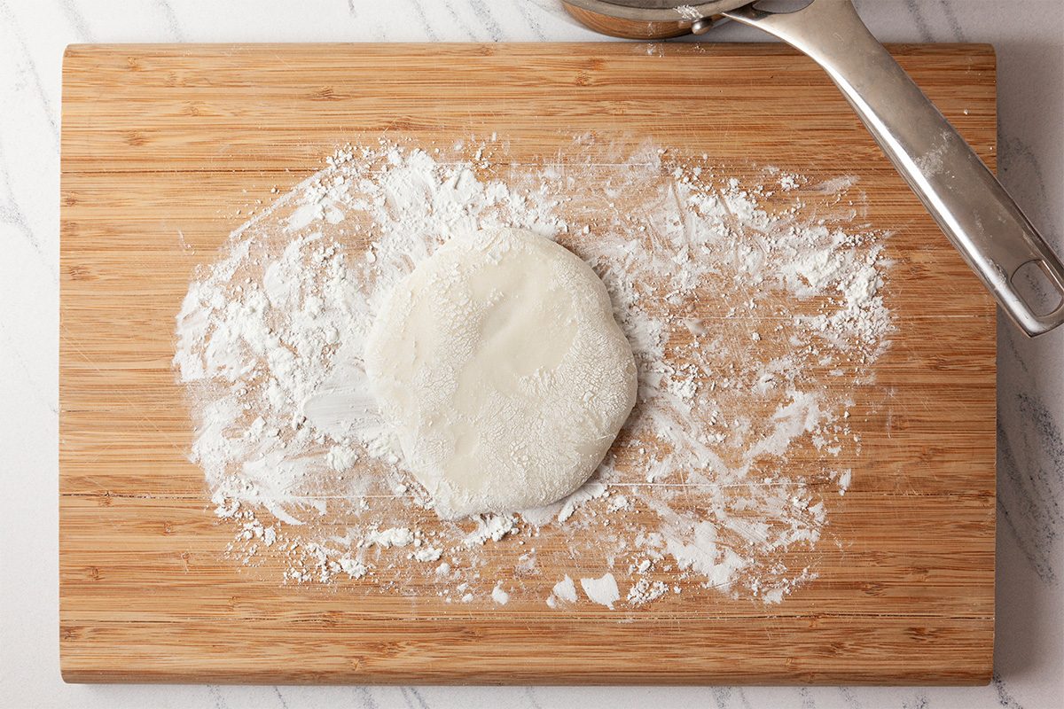 A round piece of dough sits on a wooden cutting board dusted with flour, with a metal ice cream scoop nearby.