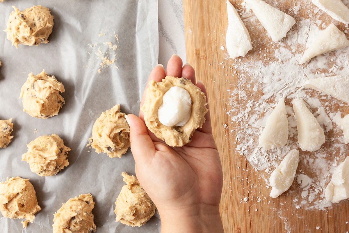 A hand holds a piece of dough with a dollop of cream in the center. On the left, more dough drops rest on parchment paper, and on the right, flour-dusted dough pieces sit on a wooden board.