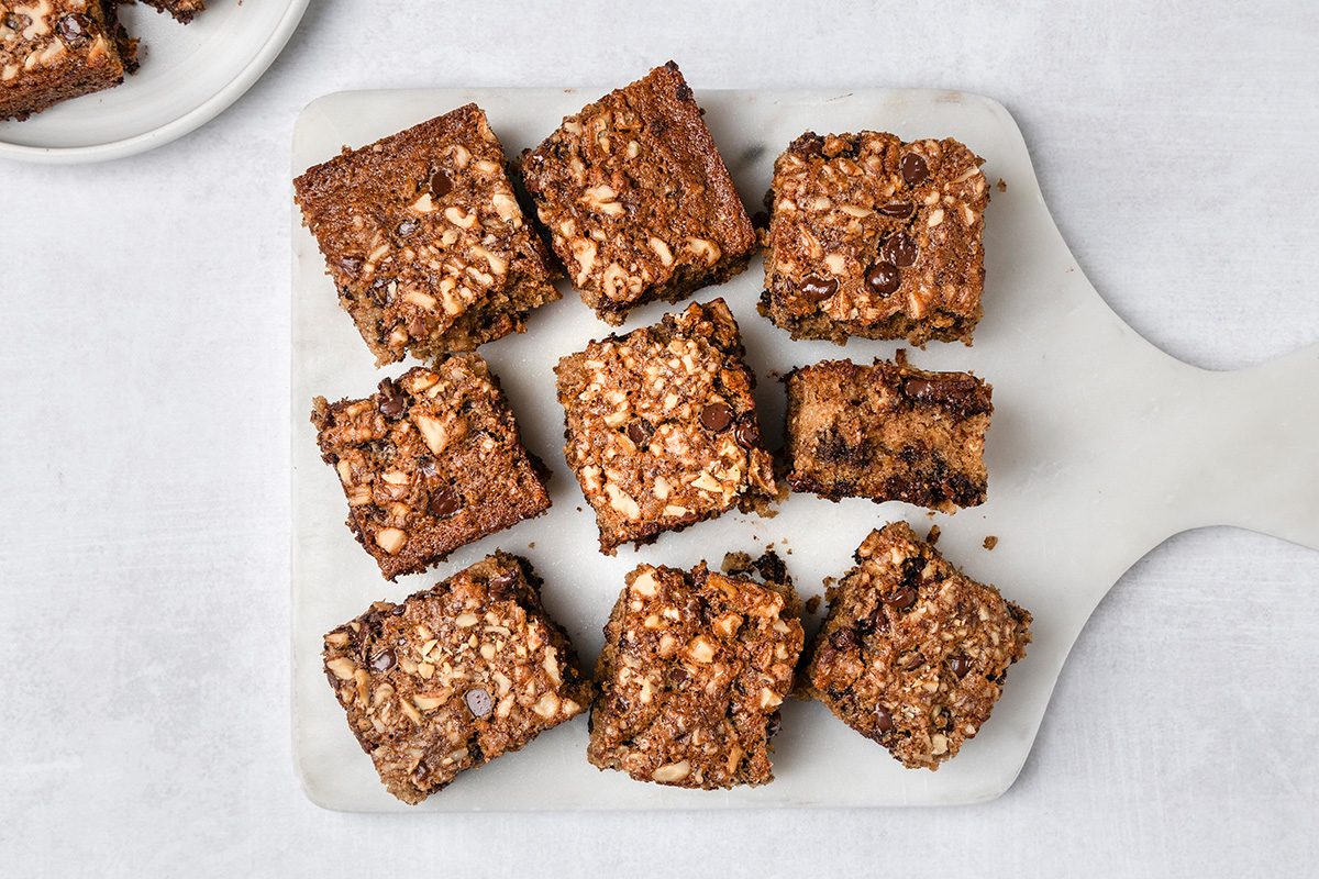 Nine square pieces of chocolate and nut brownies are arranged on a white marble serving board, with a partial view of an additional plate of brownies in the top left corner.