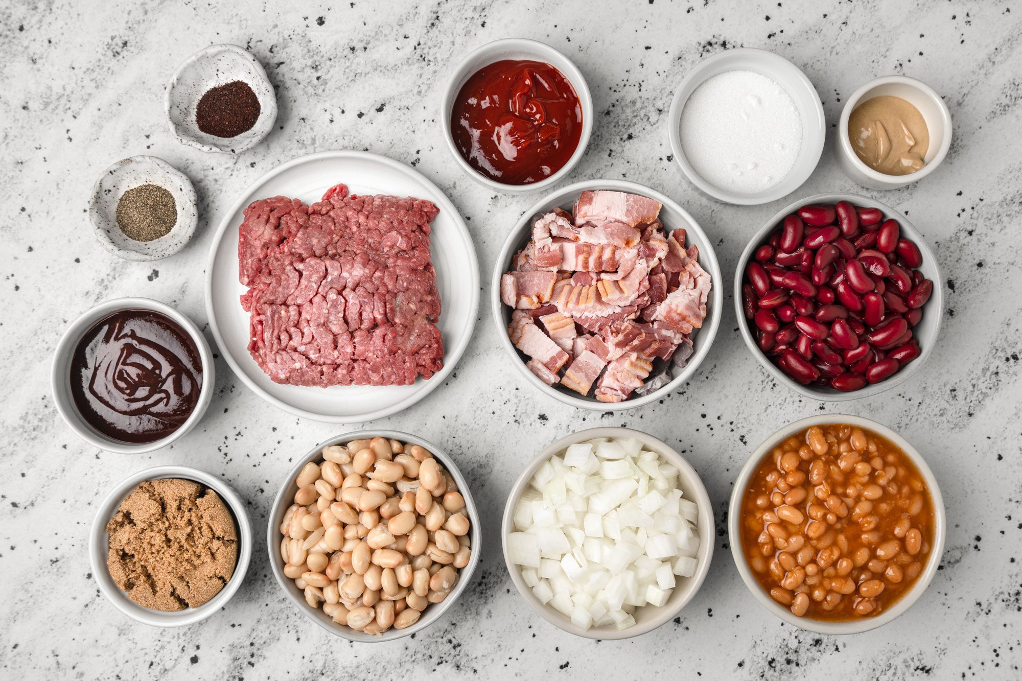 Top shot of this image shows various chili ingredients in bowls placed on a marble surface; Ingredients include ground beef; chopped bacon; kidney beans; white beans; baked beans; chopped onions; brown sugar; ketchup; barbecue sauce; spices; sugar and mustard