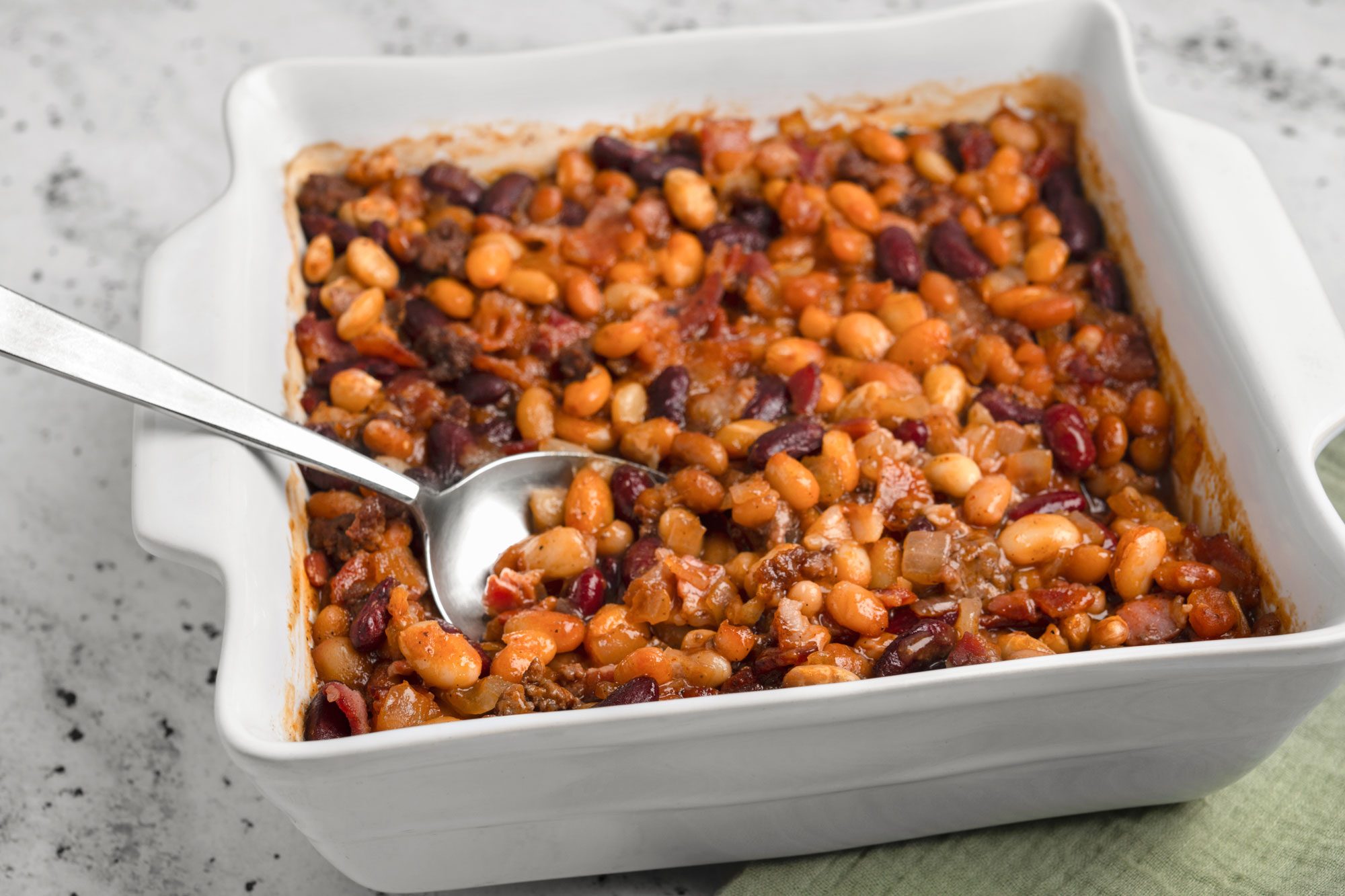 3/4th shot of a square white baking dish contains baked beans—red and white beans in a thick; reddish sauce; with a metal serving spoon resting inside. The dish is placed on a light-colored background; suggesting a homely meal setting
