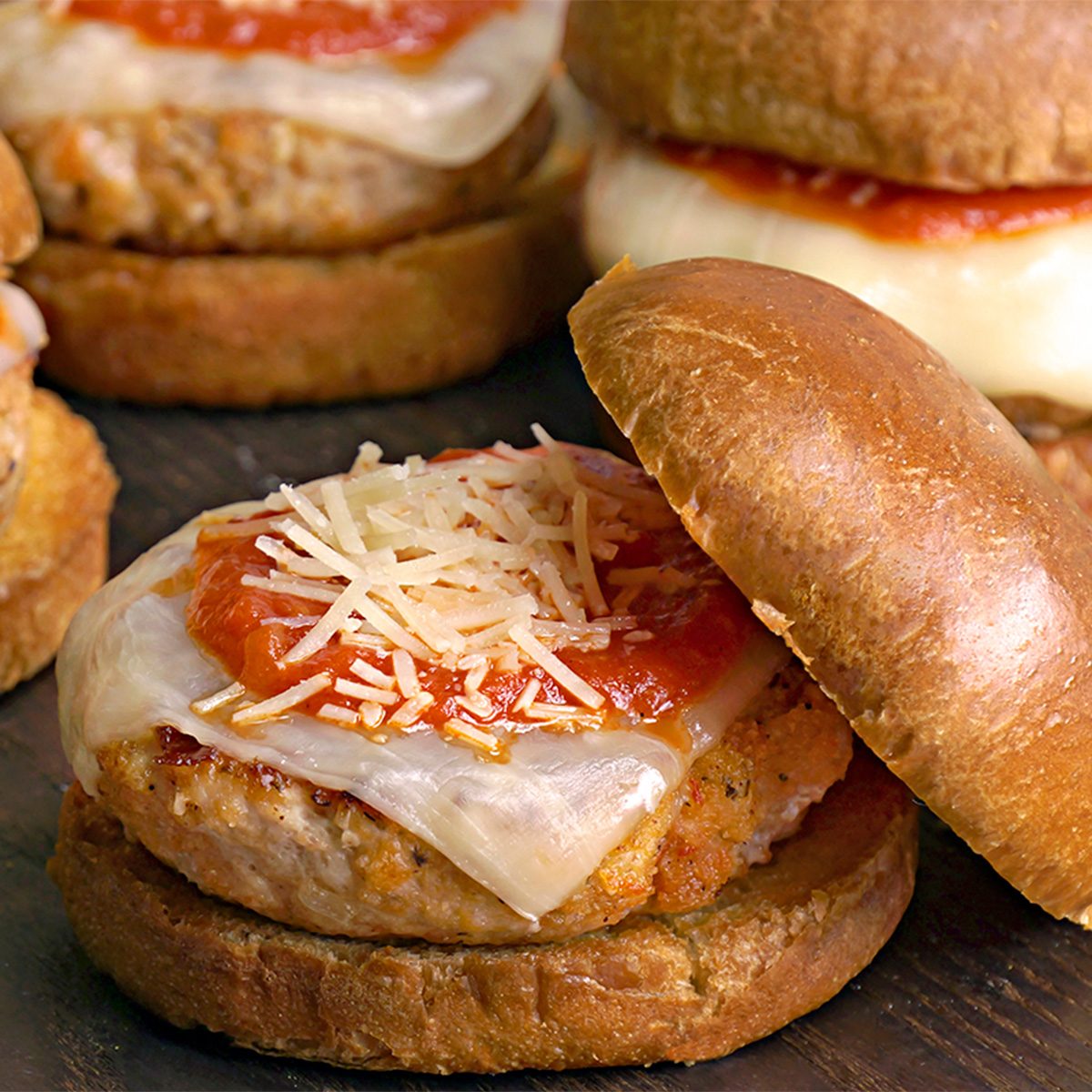 chicken parmesan sandwiches on wooden surface, each with a breaded chicken patty, melted cheese, tomato sauce, and shredded parmesan on a brioche bun. One sandwich has the top bun slightly lifted to show the ingredients.