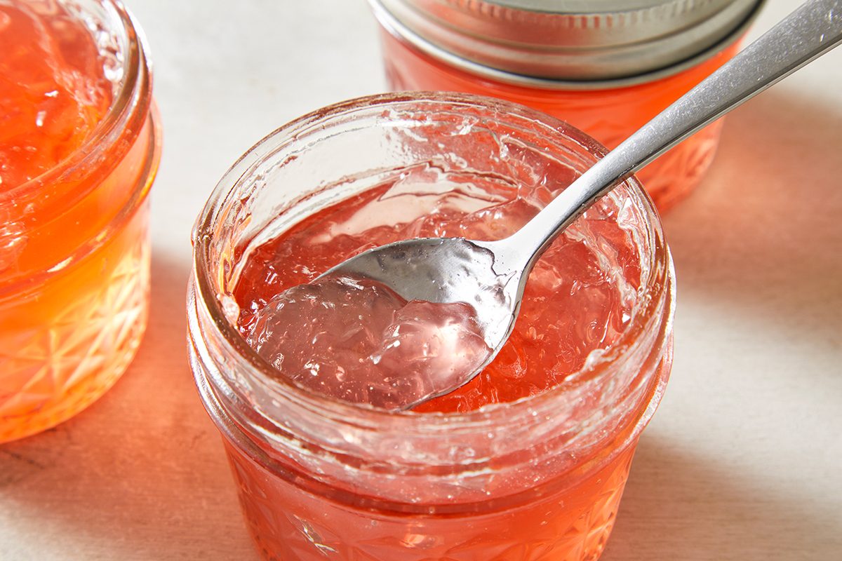 An open glass jar filled with orange-pink jelly, a spoon resting inside, with two more sealed jars of jelly in the background on a light surface.