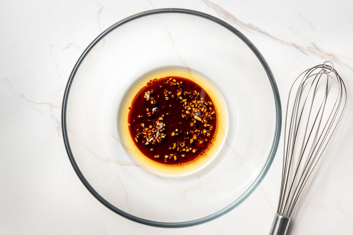 A glass mixing bowl with a reddish-brown liquid and seasoning sits on a white marble surface next to a metal whisk.