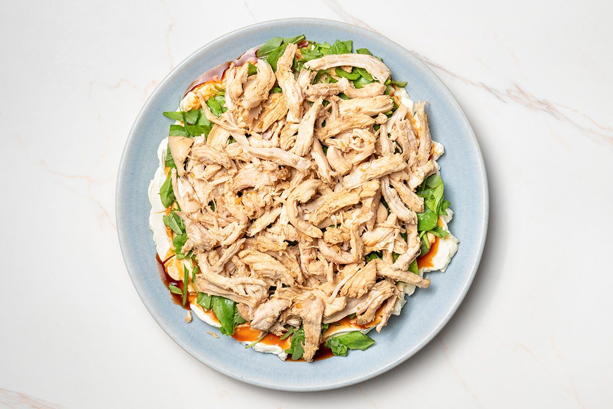 A blue plate topped with shredded cooked chicken on a bed of greens and thinly sliced vegetables, set on a white marble surface.