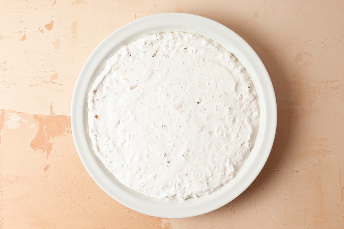 A white round dish filled with an even layer of creamy white dip, placed on a light beige surface.