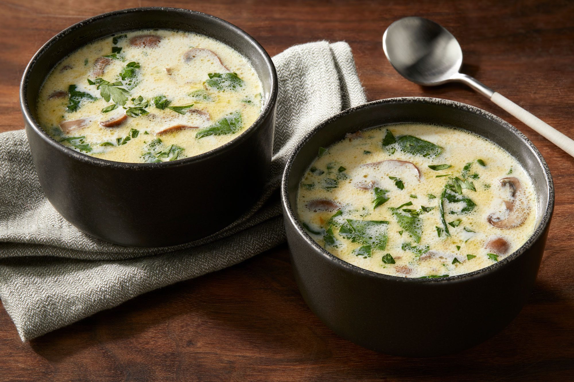 3/4 angle view shot of Creamy Cremini-Spinach Soup served in two black bowls; sprinkled with parsley; one bowl placed over a grey napkin with a spoon nearby; all set on a brown wooden surface