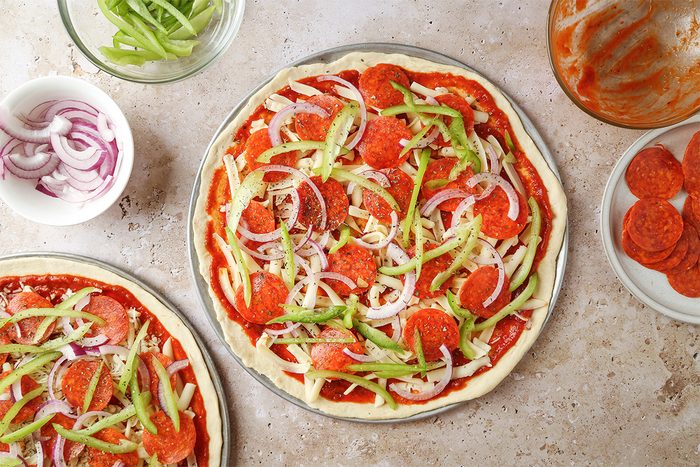 Overhead view of unbaked pepperoni pizzas topped with sliced green bell peppers and red onions on a countertop, surrounded by bowls of extra toppings and sauce.