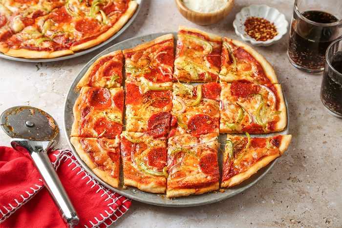 A thin-crust pepperoni pizza cut into square slices sits on a round tray, with a pizza cutter and red napkin nearby. Two glasses of soda and bowls of grated cheese and red pepper flakes are in the background.