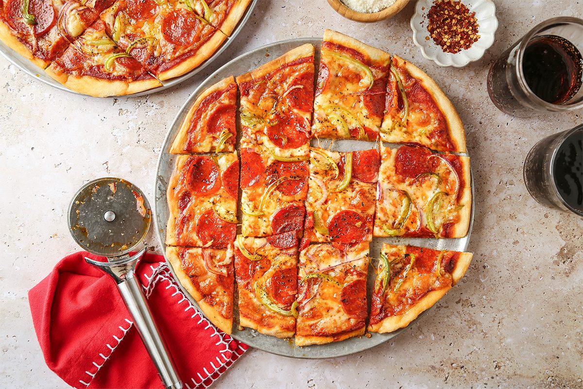Overhead view of a sliced pepperoni pizza on a round metal tray, with a pizza cutter, red napkin, bowl of grated cheese, bowl of red pepper flakes, and glasses of dark beverage on a light countertop.