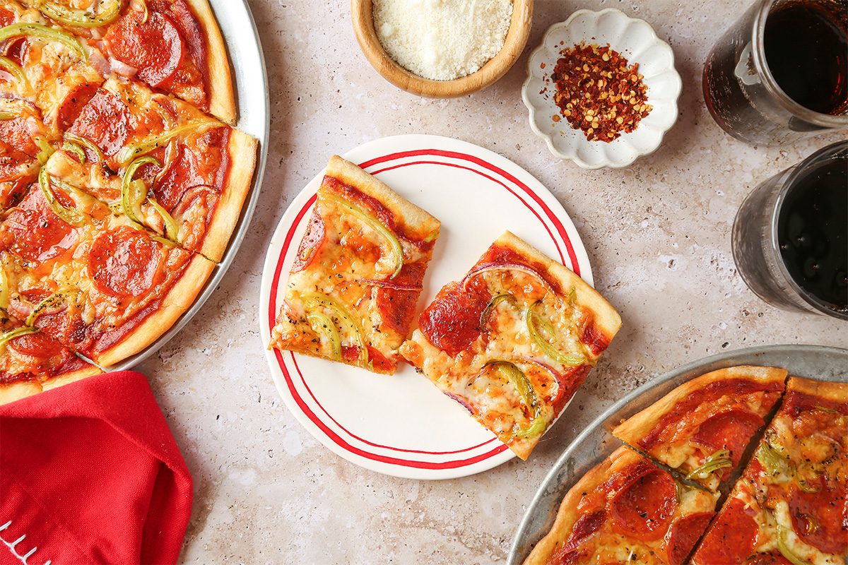 Two slices of pepperoni and green pepper pizza on a plate, surrounded by more pizza, grated cheese, red pepper flakes, drinks, and a red napkin on a light stone surface.