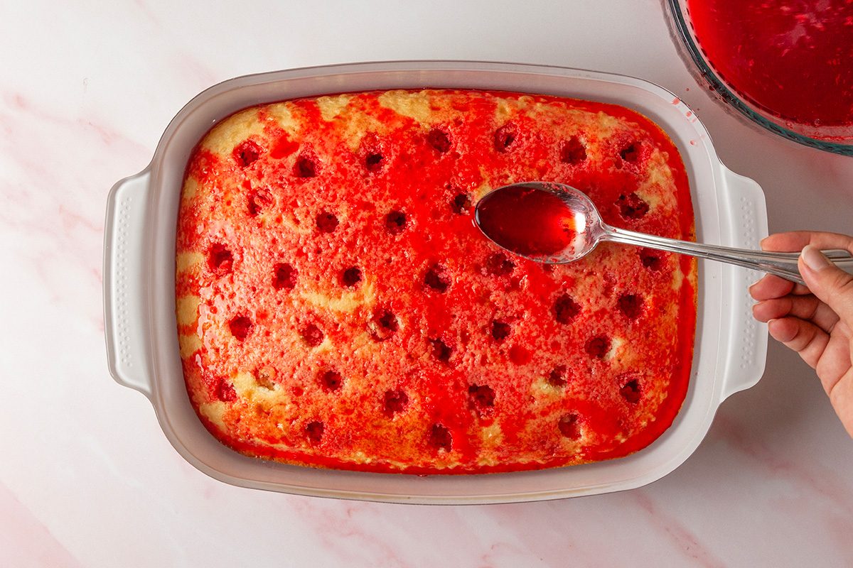 overhead shot of a hand holds a spoon, pouring red liquid into holes in a baked yellow cake in a white rectangular dish; The cake has evenly spaced holes and sits on a pink marble countertop; A bowl of red liquid is nearby