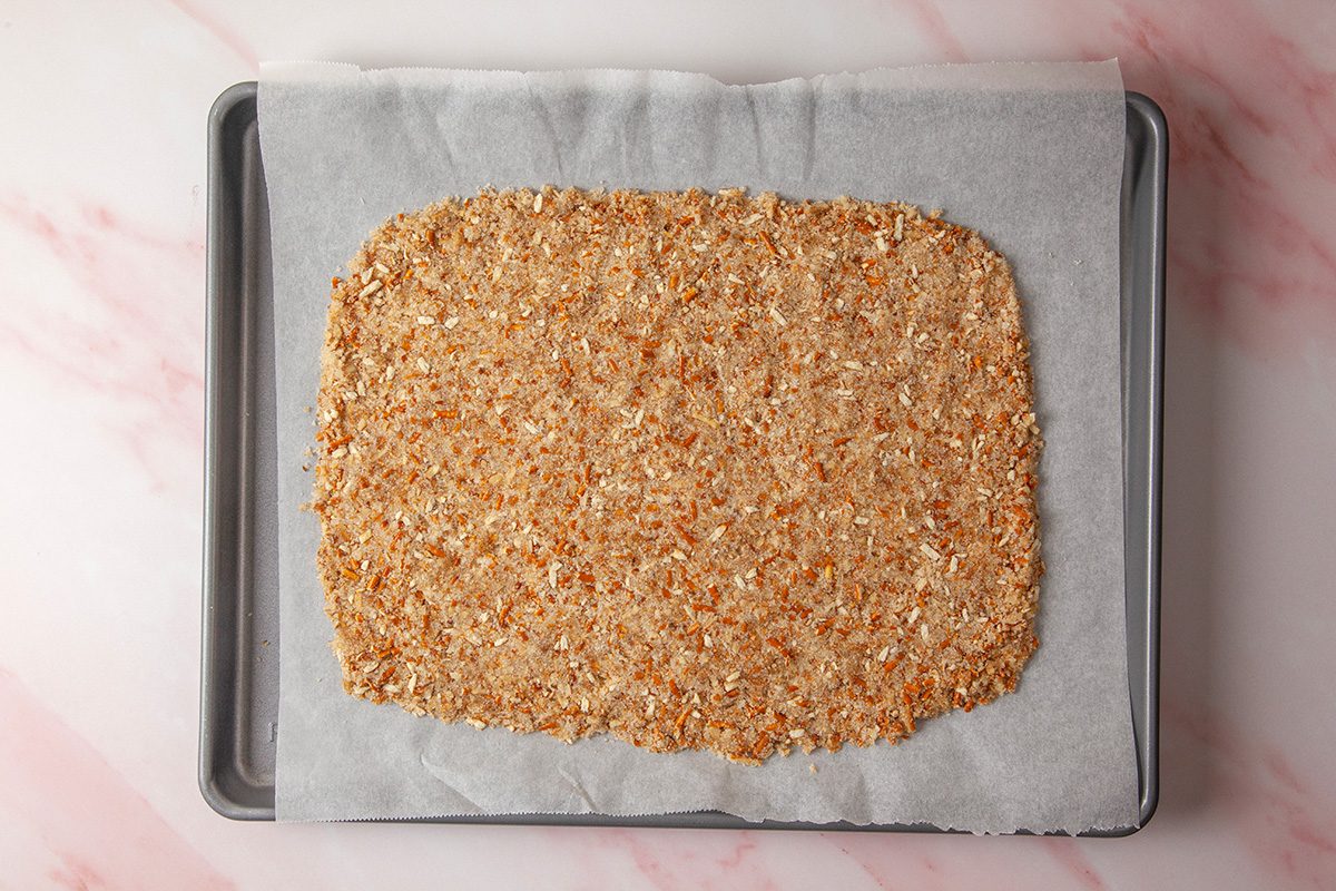 overhead shot of a rectangular sheet of uncooked cracker dough with visible oats and seeds sits on parchment paper on a baking tray, ready for baking