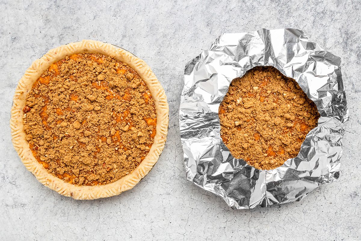 Two unbaked crumb-topped pies on a gray surface; one pie has exposed edges, while the other has its crust edges covered with crinkled aluminum foil.