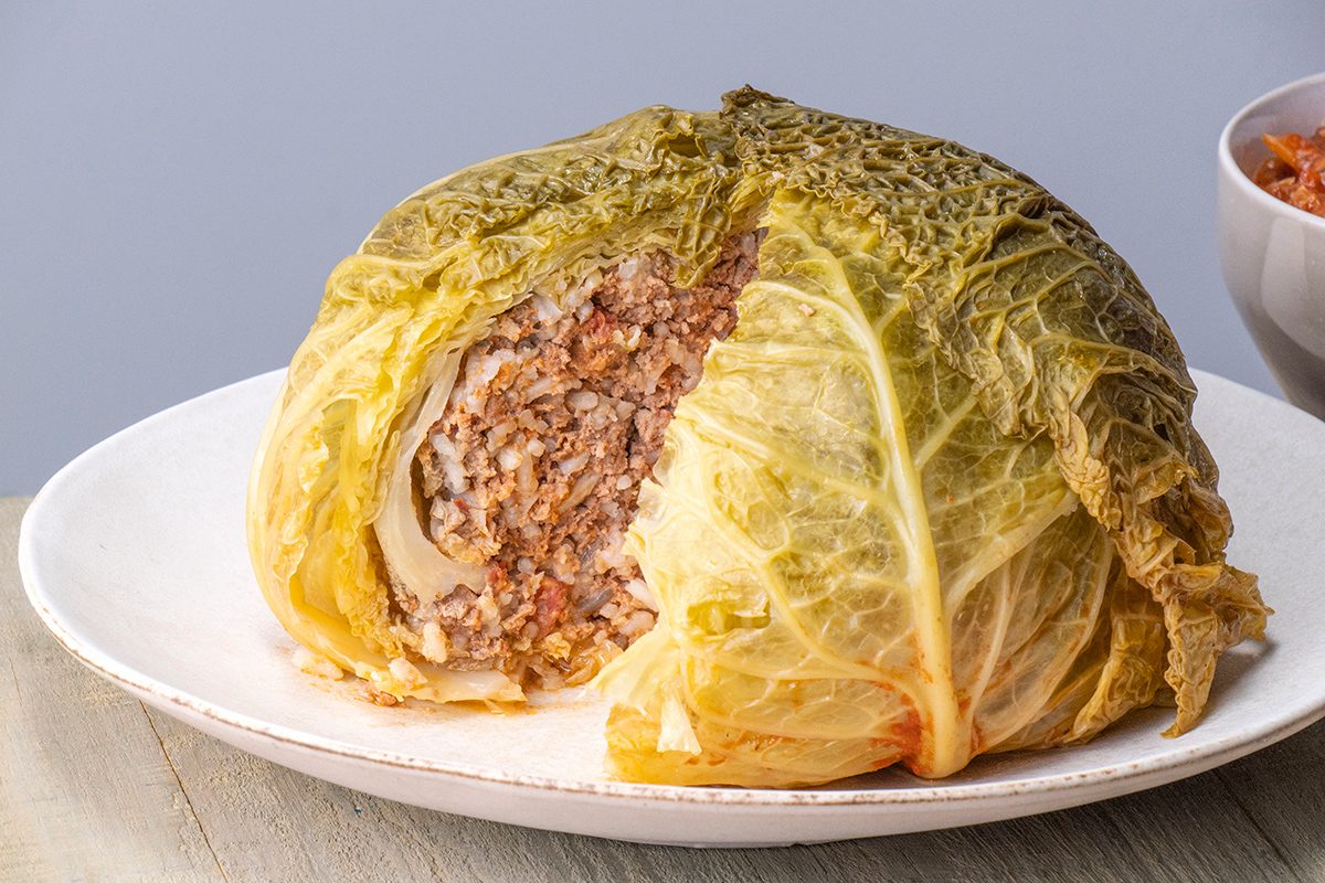 A stuffed cabbage head sits on a white plate, with a wedge cut out to reveal a filling of rice and ground meat. The cabbage leaves are cooked and slightly wilted, with the filling clearly visible inside.