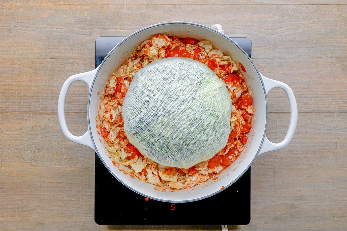 A round pot on a stovetop contains chopped cabbage and tomatoes, with a whole cabbage head placed on top of the mixture.