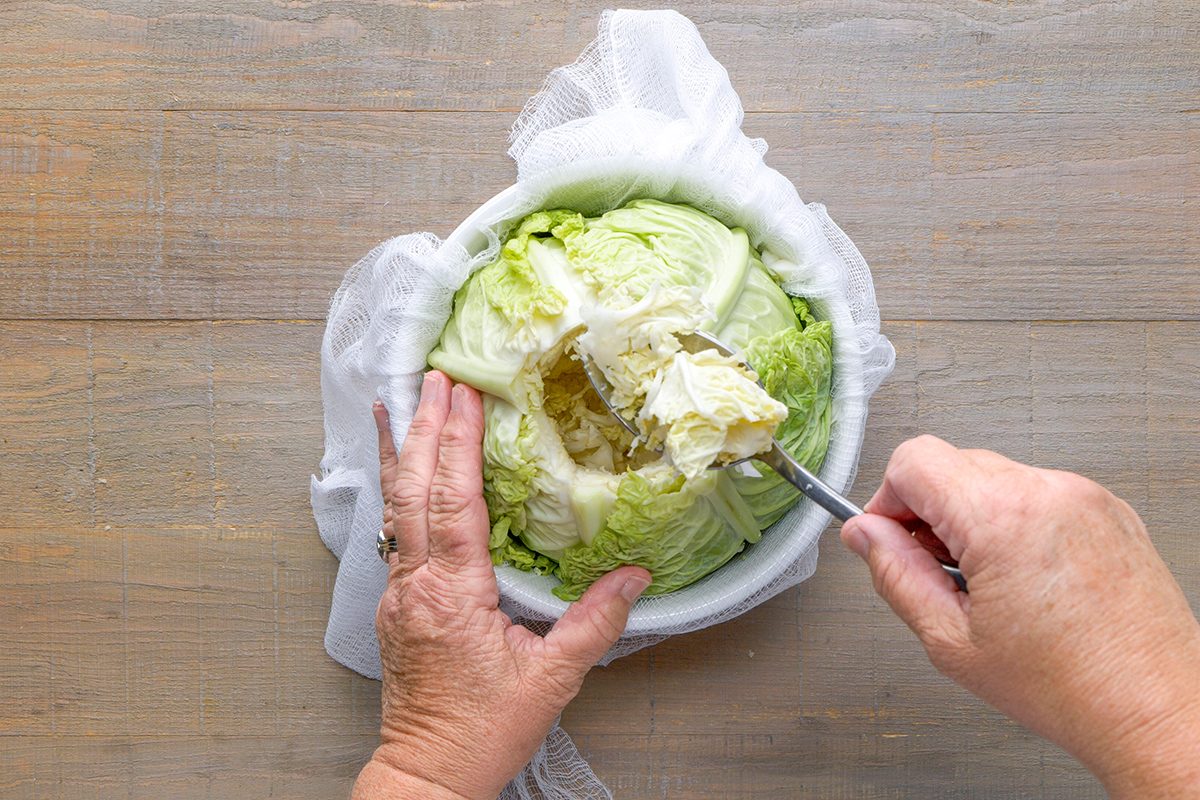 A person scooping out the center of a head of cabbage with a spoon, while holding it steady on a wooden surface covered with white cloth.