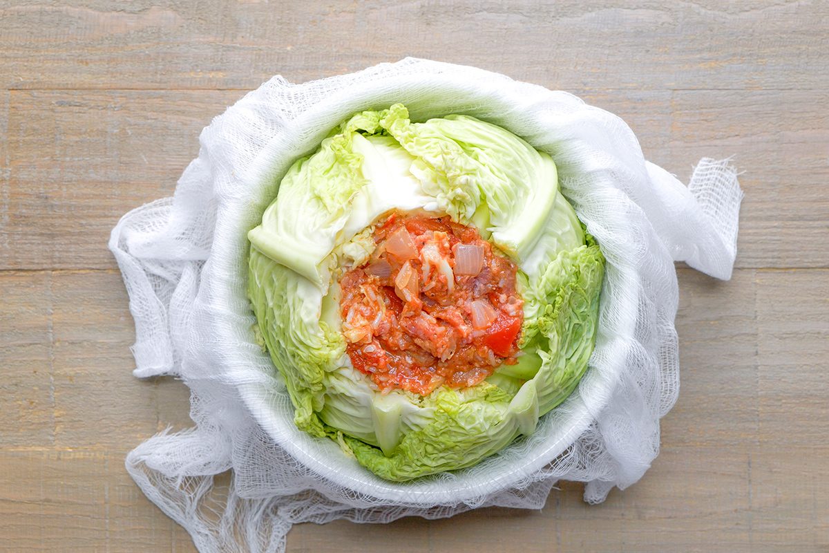 A head of napa cabbage with its center filled with diced vegetables and a red seasoning paste, placed in a white bowl lined with cheesecloth on a wooden surface.