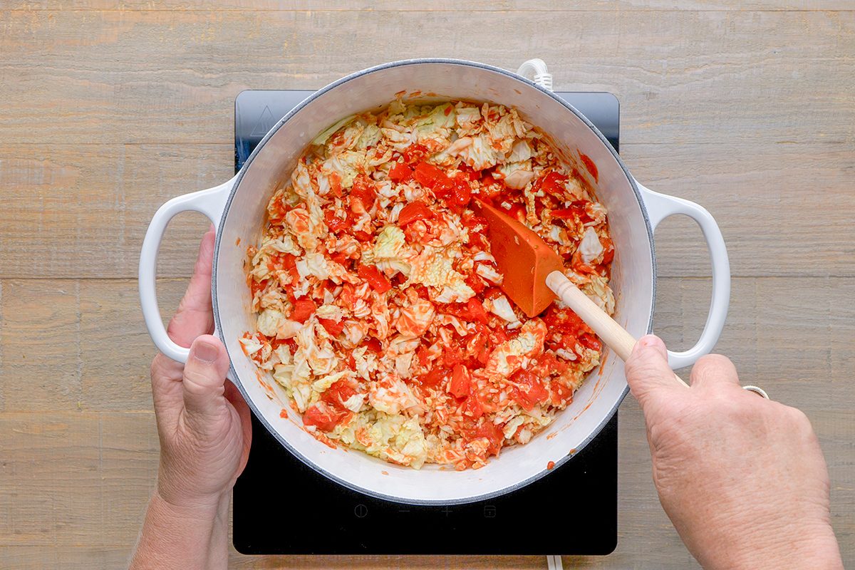 A person stirs chopped cabbage and tomatoes in a large white pot on a stovetop with a wooden spoon.